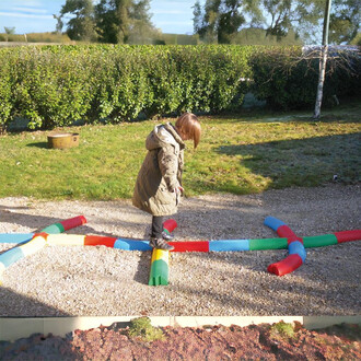 Enfant marchant sur des poutres courbes colorées disposées au sol, dans un jardin verdoyant, avec des haies en arrière-plan. Les poutres sont de différentes couleurs (rouge, vert, bleu, jaune) et créent un parcours ludique.