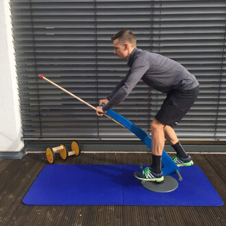 Un homme en tenue de sport utilise un équipement de jonglerie. Il est penché en avant, tenant une longue perche avec une boule rouge à son extrémité. L'équipement se trouve sur un tapis bleu, avec un axe sur lequel il se balance. À gauche, on aperçoit des