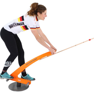 Femme souriante en tenue de cyclisme se tenant sur un appareil d'entraînement orange, avec une tige en bois, simulant un mouvement de vélo. Elle porte un maillot blanc aux couleurs de l'Allemagne et des leggings noirs. Un sol gris est visible en dessous.