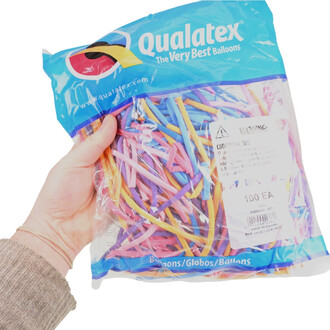 Sachet de ballons Qualatex 160Q Vibrant tenu dans une main, contenant une variété de ballons longs de couleur vive, tels que rose, bleu, violet et jaune, avec un logo Qualatex visible sur l'emballage.