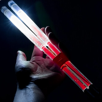 Baguettes de diabolo lumineuses tenues dans une main, avec une lumière blanche brillante à l'extrémité de chaque baguette et un détail rouge au centre, sur un fond sombre.