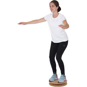 Femme en tenue de sport, debout sur un disque d'équilibre en bois, les bras tendus pour maintenir son équilibre. Elle porte un t-shirt blanc et un legging noir, avec des chaussures de sport grises et violettes.