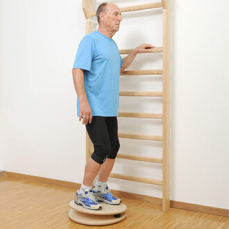 Homme debout sur une plateforme de balance, tenant une barre en bois pour équilibre, vêtu d'un t-shirt bleu et de leggings noirs, dans une pièce avec un sol en bois et un mur blanc.