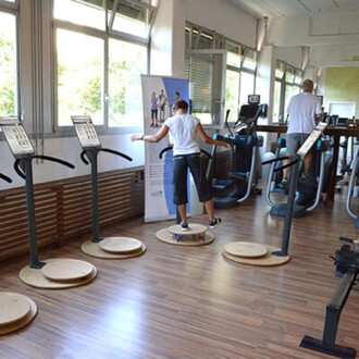 Image d'une salle de sport avec plusieurs appareils de fitness. Au premier plan, une personne se tient sur une plateforme de balance, les bras écartés pour garder l'équilibre. À l'arrière-plan, on peut voir deux autres personnes utilisant des machines de 