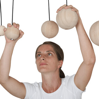 Femme en t-shirt blanc tenant deux balles en bois suspendues par des cordes, regardant vers le haut avec concentration.