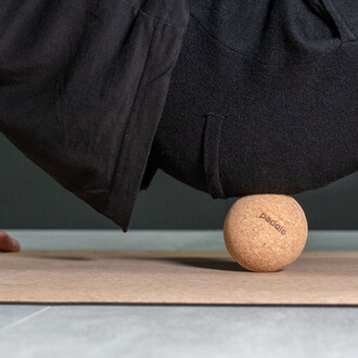 Balle en liège de massage sur un tapis, avec un pied en position de déséquilibre au-dessus. Le vêtement est sombre, soulignant la tension de l'exercice.