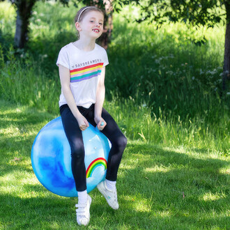 Enfant sautant sur le ballon sauteur Tobar 60cm dans un espace vert, portant un t-shirt à motif arc-en-ciel.