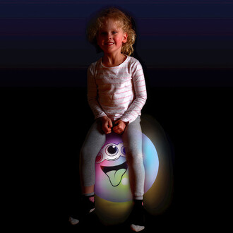 Jeune fille assise sur un ballon sauteur lumineux translucide de taille junior avec des lumières LED activées par mouvement. Le ballon présente un visage souriant et est conçu pour les enfants de 5 ans et plus. Parfait pour le développement de la motricit