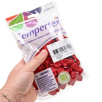 Sac transparent contenant des ballons rouges en forme de cœur, tenu par une main ; emballage Sempertex avec logo, étiquettes et code-barres visibles.
