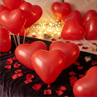 Ballons en forme de cœur rouges gonflés, décor romantique avec pétales rouges et guirlande lumineuse floue en arrière-plan sur un drap noir.