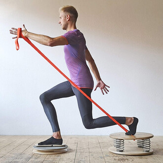 Homme en train de s'exercer sur une plateforme de balance avec des ressorts, utilisant une bande élastique rouge. Il est vêtu d'un t-shirt violet et de leggings noirs, en position de course, avec une jambe en avant et les bras tendus pour stabiliser son c