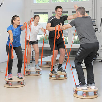 Quatre personnes s'entraînent en utilisant des équipements de fitness sur des plateformes circulaires avec des ressorts. Elles tiennent des bandes élastiques orange, en position dynamique dans une salle lumineuse.