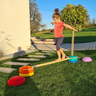 Enfant en équilibre sur une barre, pieds nus, dans un jardin verdoyant. La barre est placée sur des bases colorées (rouge, orange, bleu et violet) et le soleil brille, créant une ombre sur l'herbe.