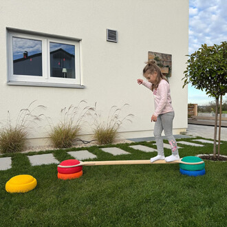 Une petite fille marche sur une barre d'équilibre en bois, placée entre des dispositifs ronds colorés (rouge, jaune, orange, bleu et vert) sur une pelouse verte. En arrière-plan, on aperçoit une maison blanche avec des fenêtres et un petit arbre. Un ciel 