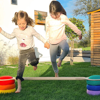 Deux jeunes filles sautent joyeusement sur une barre d'équilibre en bois, main dans la main. Elles sont en extérieur, sur une pelouse verte, avec une ambiance ensoleillée. À côté d'elles, un empilement de disques colorés (rouge, orange, jaune, vert, viole