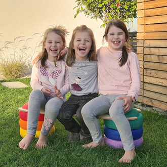 Trois filles assises sur un empilement de sièges colorés, riant et souriant. Elles portent des vêtements confortables : une shirt à manches longues avec des motifs, un t-shirt gris avec une illustration et un pull rose à pois. Elles sont en extérieur, sur