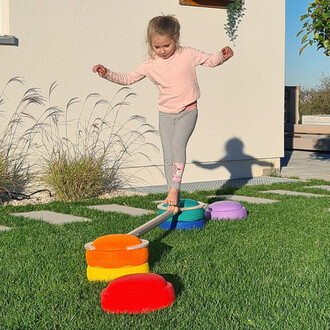 Petite fille en équilibre sur une barre, avançant sur des coussins colorés disposés sur l'herbe. Elle porte un pull à petits pois et des leggings gris, avec une posture concentrée et les bras tendus pour maintenir son équilibre. En arrière-plan, une maiso