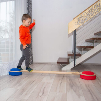 Un enfant en sweat à capuche orange marche sur une barre d'équilibre en bois, posée sur deux supports en forme de coussins, un bleu et un rouge. L'arrière-plan montre un intérieur moderne avec des escaliers et des rideaux.