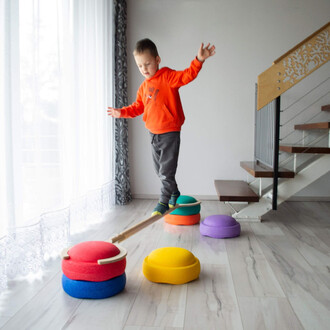 Enfant en sweatshirt orange marchant sur une barre d'équilibre, avec des éléments colorés en dessous (rouge, bleu, vert, orange, violet, jaune) dans un intérieur lumineux avec des rideaux. Un escalier en bois est visible à droite.