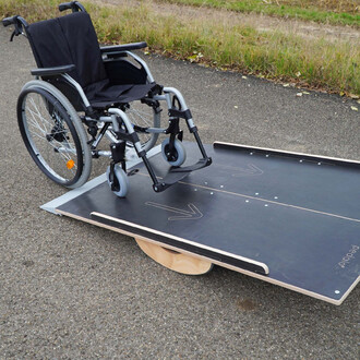 Image d'une rampe pour fauteuil roulant avec un fauteuil roulant placé dessus, sur une surface routière, entourée d'herbe. La rampe est en bois, avec des flèches marquées pour indiquer la direction.