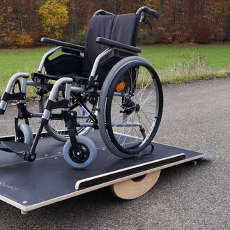 Image d'un fauteuil roulant posé sur une plateforme stable avec une grande roue à l'arrière, sur une surface asphaltée et entourée d'un paysage naturel avec des arbres aux couleurs d'automne.