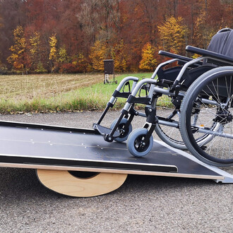 Rampe d'accès pour fauteuil roulant avec un fauteuil roulant surmontant une surface noire, posée sur un sol en bitume. En arrière-plan, un paysage d'automne avec des arbres aux couleurs chaudes.