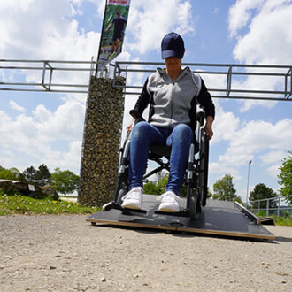 Femme en fauteuil roulant utilisant une rampe pour monter sur une surface surélevée, avec un ciel partiellement nuageux en arrière-plan et une colonne de pierre à proximité.