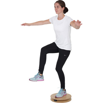 Femme en tenue de sport, exécutant un exercice d'équilibre sur une planche rotative. Elle est debout sur une jambe, l'autre genou relevé, bras étendus pour maintenir son équilibre. Elle porte un t-shirt blanc et un leggings noir avec des chaussures de spo