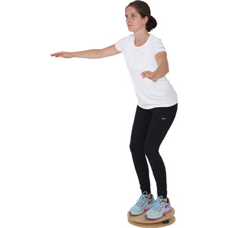 Femme en équilibre sur un disque de stabilité, les bras tendus sur le côté pour maintenir sa posture. Elle porte un t-shirt blanc et des leggings noirs, avec des chaussures de sport aux teintes bleu et violet. Fond blanc neutre.