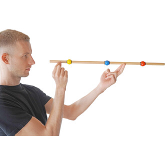 Un homme en t-shirt noir tient une barre en bois avec trois balles colorées (jaune, bleue et rouge) positionnées dessus. Il pointe avec un doigt l'une des balles, tandis que l'autre main est utilisée pour maintenir la barre. L'arrière-plan est blanc.