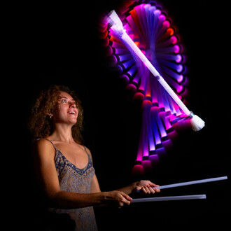 Une personne souriante jongle avec un bâton du diable lumineux, levé dans les airs, tandis que des motifs colorés se forment autour. La scène est sous un fond noir, mettant en valeur les lumières vives du bâton et l'expression joyeuse du jongleur.