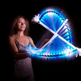 Une personne souriante utilise un bâton du diable lumineux dans une pièce sombre, créant des effets de lumière vibrants avec des mouvements fluides. Les bâtons sont éclairés de bleu et de blanc, générant une belle traînée lumineuse. La personne porte une