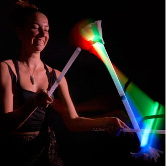 Une personne souriante jongle avec des bâtons du diable lumineux de couleur, créant des effets lumineux multicolores. L'arrière-plan est sombre, mettant en valeur les mouvements et lumières des bâtons. Des bandes de lumière verte, rouge et bleue émergent