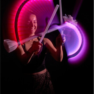 Une personne souriante jongle avec deux bâtons lumineux, créant des effets de lumière colorés dans un environnement sombre. Les bâtons sont en mouvement, produisant des halos de lumière rose et violet.