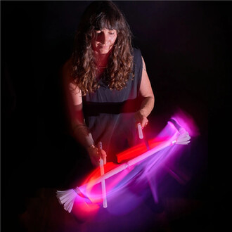 Femme jonglant avec un bâton du diable lumineux, entourée de lumières colorées en mouvement, sur fond noir. Ses cheveux longs et ondulés encadrent son visage concentré. Le bâton du diable se compose de deux extrémités coniques blanches et de bâtonnets lum