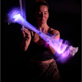 Femme manipulant un bâton du diable lumineux avec des embouts en plastique, créant des effets de lumière colorés dans un environnement sombre. Elle sourit tout en effectuant des mouvements avec le bâton, mettant en avant la fluidité et le dynamisme de la