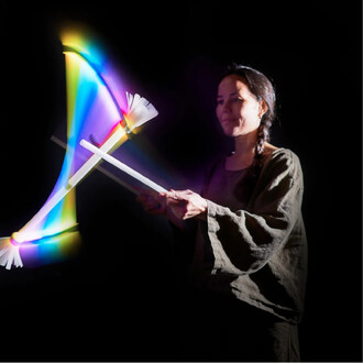 Jeune femme jonglant avec un bâton du diable lumineux, produisant des effets de lumière colorés dans un environnement sombre. Elle tient deux bâtons blancs, avec des pompons au bout, pendant qu'elle exécute des mouvements gracieux.