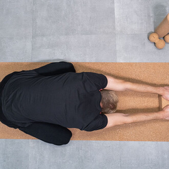 Homme en position de stretching allongé sur un tapis en liège, les bras étendus devant lui, dans un environnement intérieur avec un sol en carrelage.