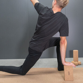 Homme en position de yoga, vêtu d'un t-shirt noir et de leggings noirs, utilisant un bloc en liège sur un tapis. En arrière-plan, plusieurs blocs de liège et balles de jonglage. Mur gris.