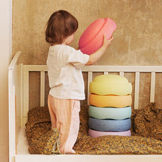 Un enfant se tient dans un lit avec un matelas recouvert de draps colorés. Il soulève un gros cercle en mousse rose au-dessus de sa tête. À côté de lui, une pile de cercles en mousse de différentes couleurs (jaune, orange, bleu, vert, lavande) est empilée