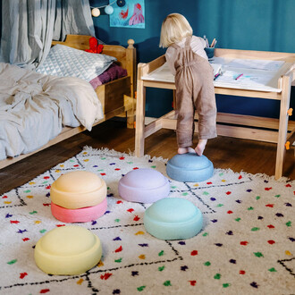 Un enfant se tient debout sur un coussin coloré dans une chambre, entouré de plusieurs coussins ronds aux couleurs pastel (jaune, rose, violet et bleu) sur un tapis blanc avec des motifs colorés. Un bureau en bois est visible à côté de l'enfant, qui regar