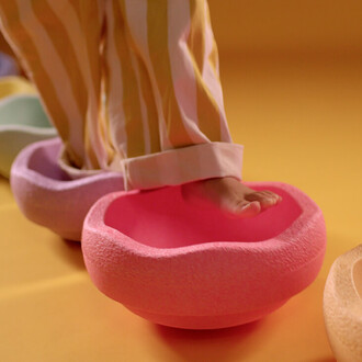 Pied d'un enfant posant sur un accessoire de jonglerie coloré en forme de bol. Le bol est rose, entouré d'autres bols de différentes couleurs (vert, violet, jaune) sur un fond jaune. L'enfant porte un pantalon à rayures.