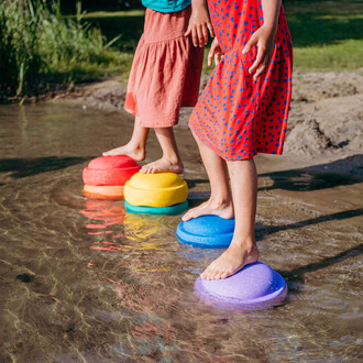 Trois enfants se tiennent en équilibre sur des disques colorés (rouge, jaune, vert, bleu et violet) posés sur un plan d'eau peu profond. Les enfants sont pieds nus, et l'on voit des reflets de lumière sur la surface de l'eau. Ils portent des vêtements est