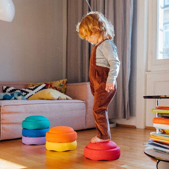 Un enfant avec des cheveux bouclés se tient debout sur un coussin de couleur rouge, dans un salon lumineux. À côté, des coussins de différentes couleurs (vert, bleu, orange et jaune) sont empilés. En arrière-plan, un canapé rose et des coussins décoratifs