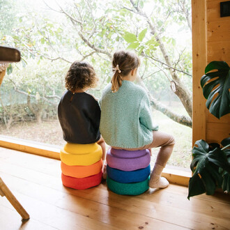 Deux enfants assis sur des coussins colorés, regardant par une grande fenêtre. L'un porte un pull en laine bleu et l'autre un vêtement noir. La pièce est lumineuse et entourée de verdure.