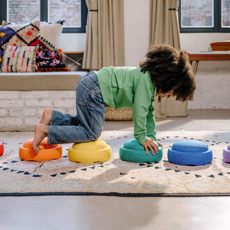 Enfant jouant sur des éléments de jonglerie colorés, avec un pull vert et des jeans, en équilibre sur des blocs dans un espace intérieur chaleureux, avec un tapis et des coussins en arrière-plan.