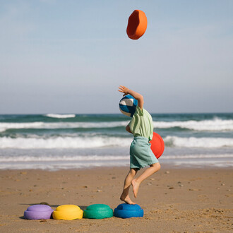 Un enfant en maillot de bain sur la plage, en train de jongler avec des balles colorées tout en sautant sur des disques en mousse. En arrière-plan, on aperçoit la mer et des vagues.