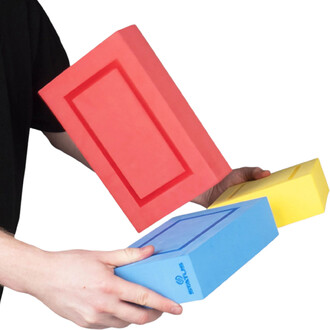 Trois blocs rectangulaires en mousse colorée (rouge, bleu et jaune) tenus par des mains, chaque bloc présentant un cadre en relief et le logo Status sur le bloc bleu.