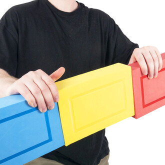 Homme en t-shirt noir tenant une longue boîte en mousse tricolore composée de trois blocs bleu, jaune et rouge reliés, chacun orné d’un motif rectangulaire.