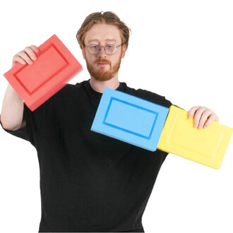 Homme avec lunettes tenant trois blocs rectangulaires en mousse colorés (rouge, bleu et jaune), style boîtes cigare Status.